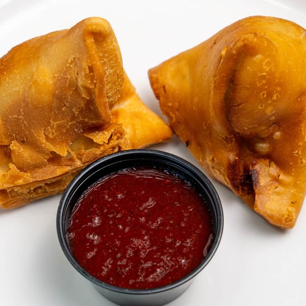 Two golden-brown samosas with a small black cup of red dipping sauce on a white surface.