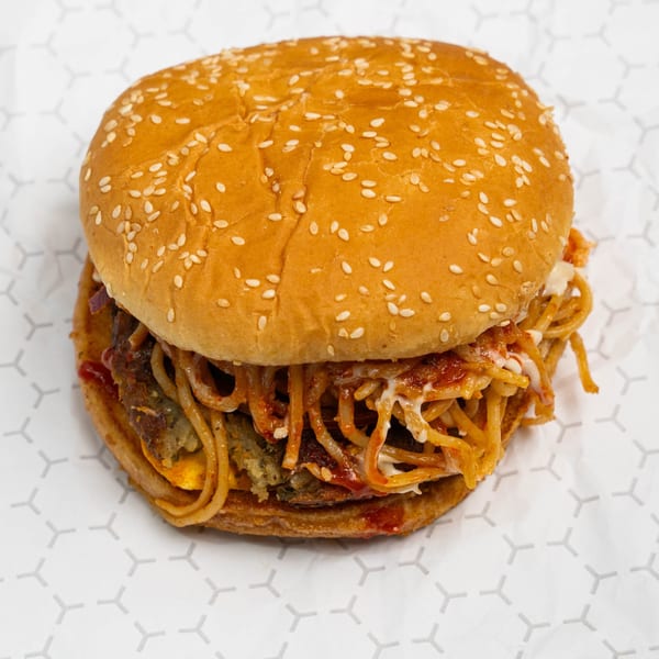 A burger with a sesame seed bun, beef patty, cheese, and spaghetti noodles on white paper.